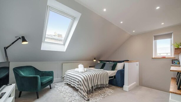 A bedroom with a skylight, a bed with a striped blanket, a green chair, and a desk with a lamp.