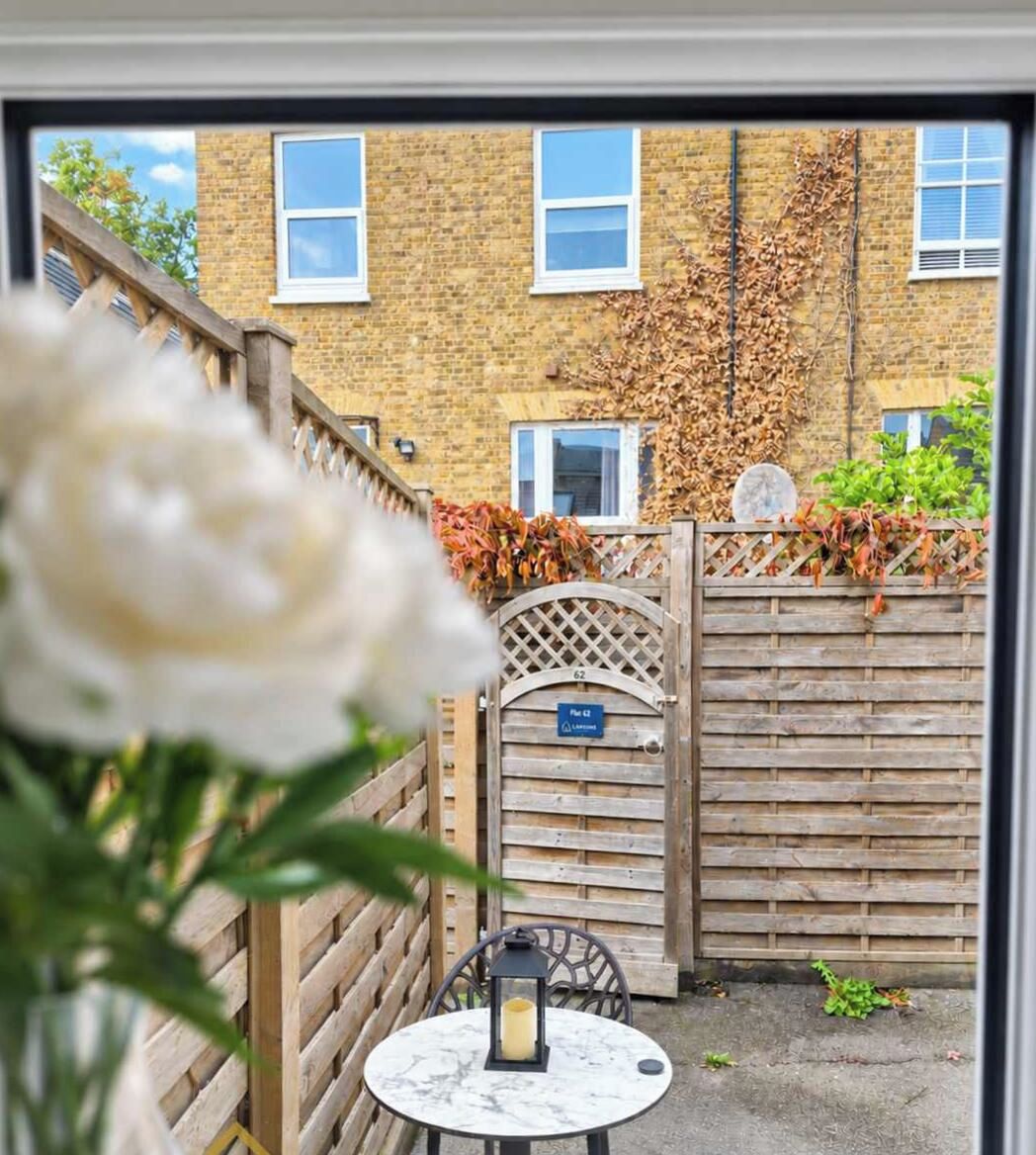 A view through a window of a courtyard with a wooden gate, a table with a lantern, and a brick building with windows in the background.