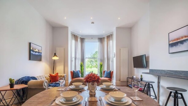 A modern dining area with a table set for four, a sofa with cushions, a TV mounted on the wall, and a window with curtains. - 3-Bed, 3-Bath Apartment with Southsea Common Views (Portsmouth)