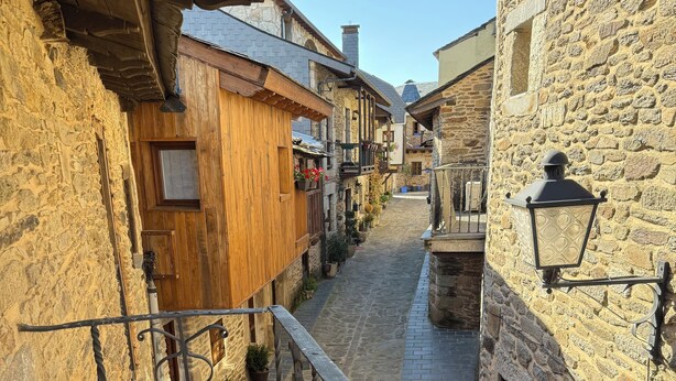 Un estrecho callejón empedrado flanqueado por edificios de piedra tradicionales con balcones de madera y barandillas de hierro forjado.