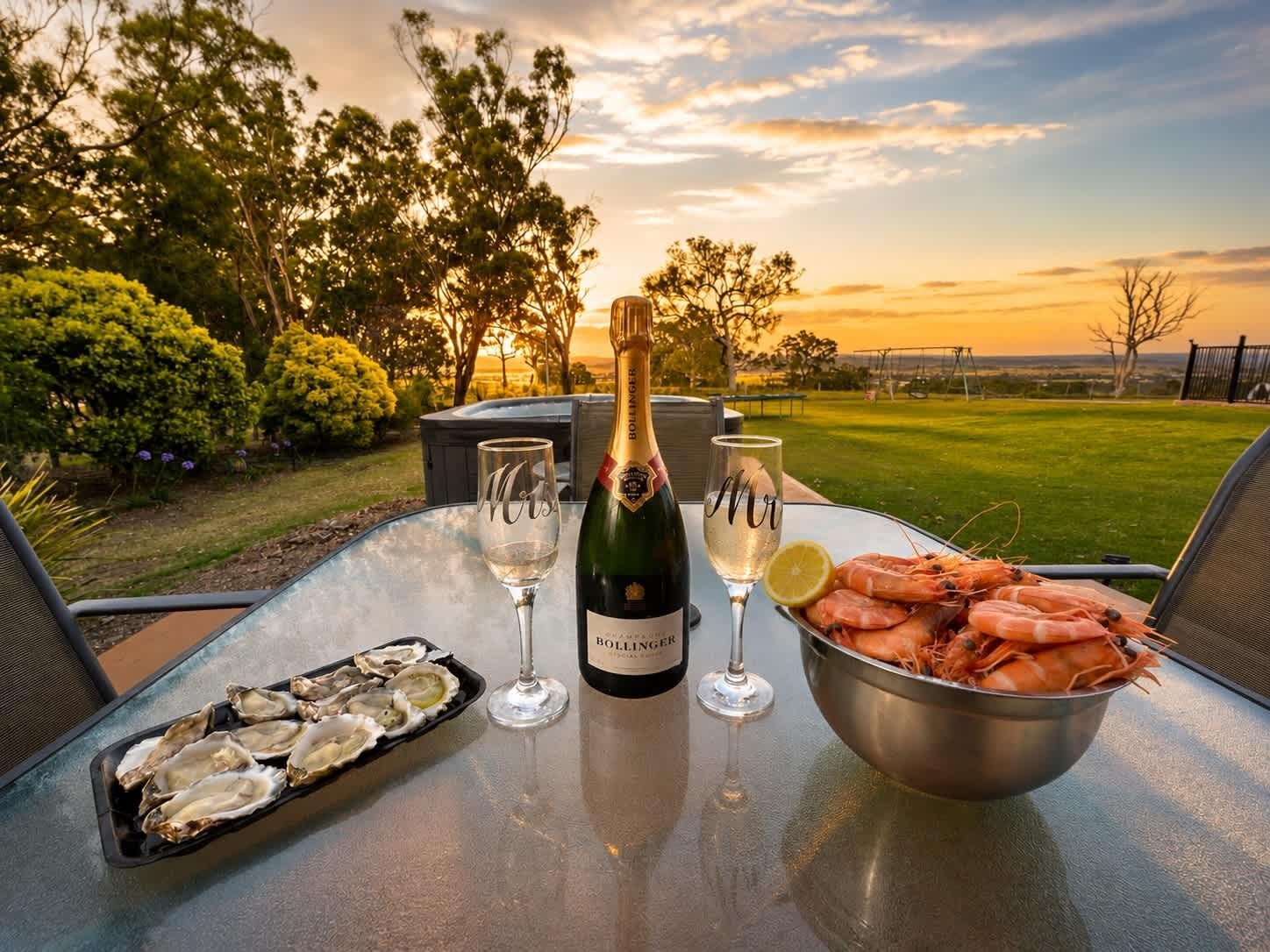 An outdoor dining table with a bottle of champagne, two glasses, a bowl of shrimp, and a tray of oysters.