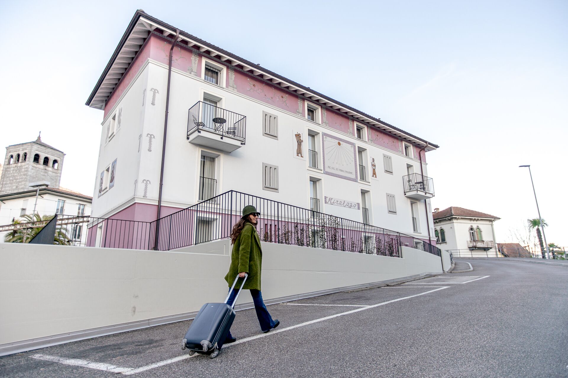 A person walking with a suitcase on a street with a building that has balconies and a sign reading "Rialaga".