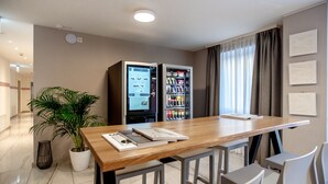 Une chambre d’hôtel moderne avec une table à manger en bois, deux tabourets de bar, une plante en pot, un mini-bar et une grande fenêtre avec des rideaux.