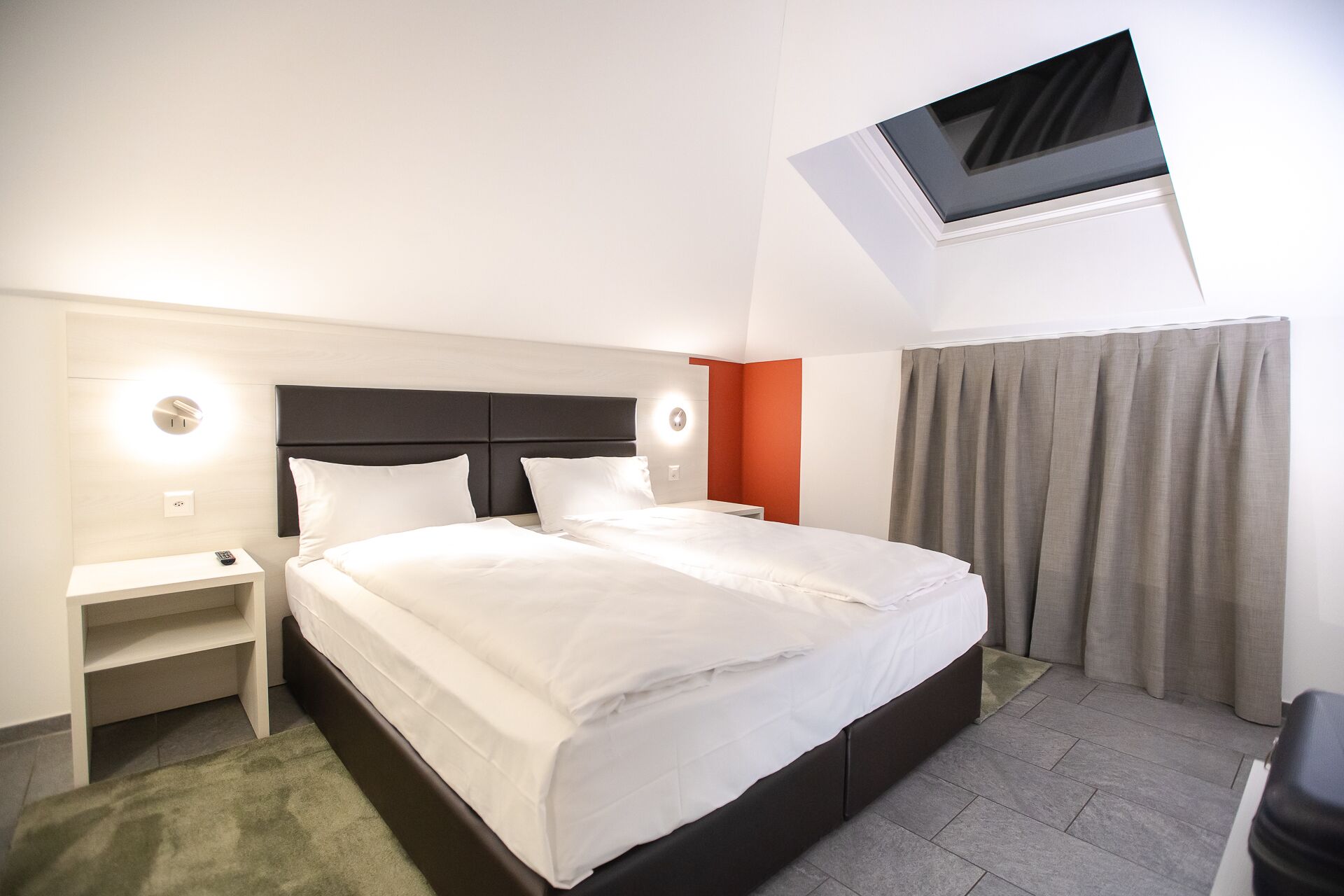 A hotel room with a large bed, a bedside table, a nightstand, a TV mounted on the wall, and a window with curtains.