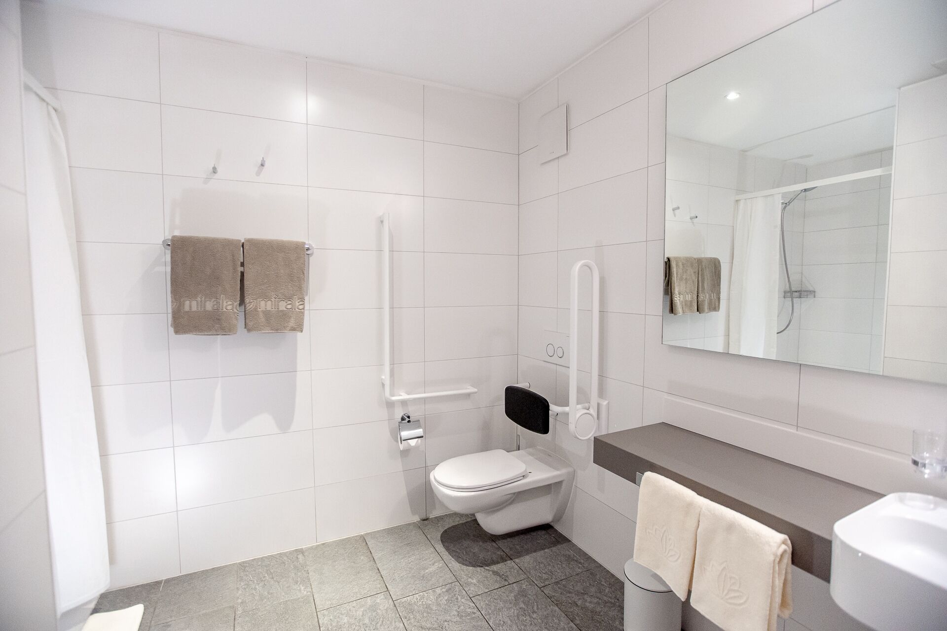 Een moderne badkamer met witte tegels, een toilet, een douche met handdouche, een handdoekrek en een grote spiegel.