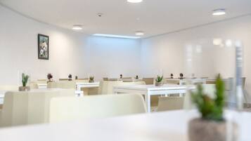 Une salle de conférence moderne et bien éclairée, avec des tables et des chaises blanches, dotée de luminaires encastrés au plafond et d’une grande fenêtre.