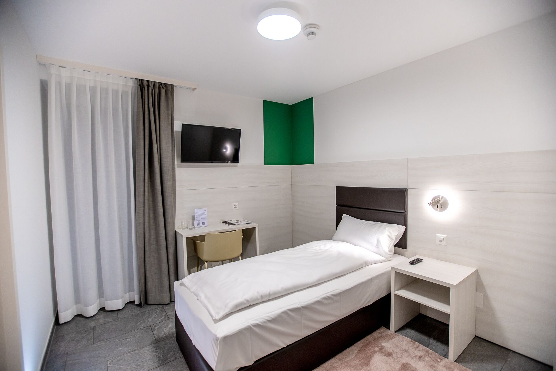 Ein Hotelzimmer mit Bett, Nachttisch, Schreibtisch mit Stuhl, einem an der Wand befestigten Fernseher und einer grünen Akzentwand.