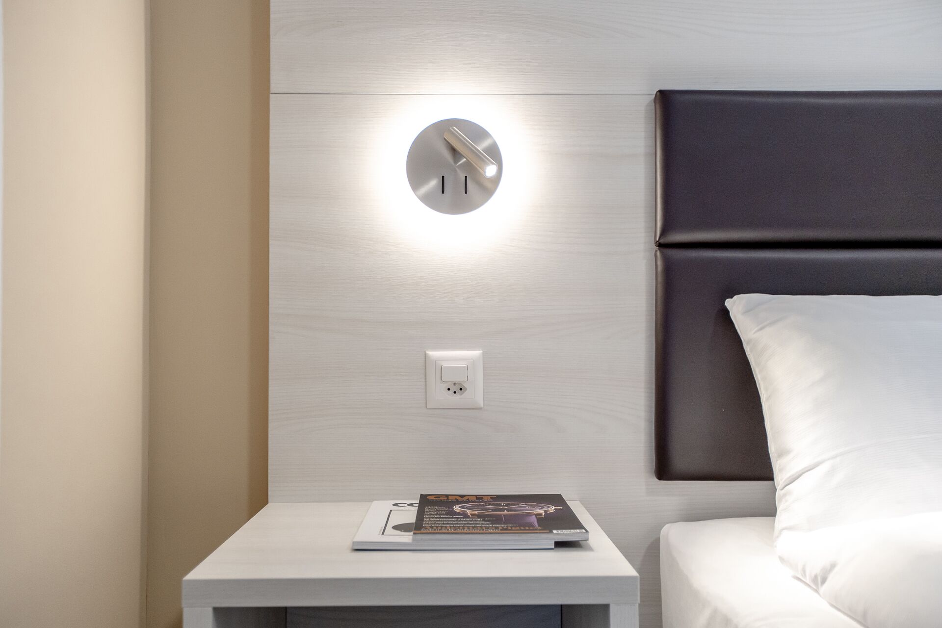 A modern hotel room with a minimalist design, featuring a bedside table with a magazine, a wall-mounted light, and a bed with a dark headboard.