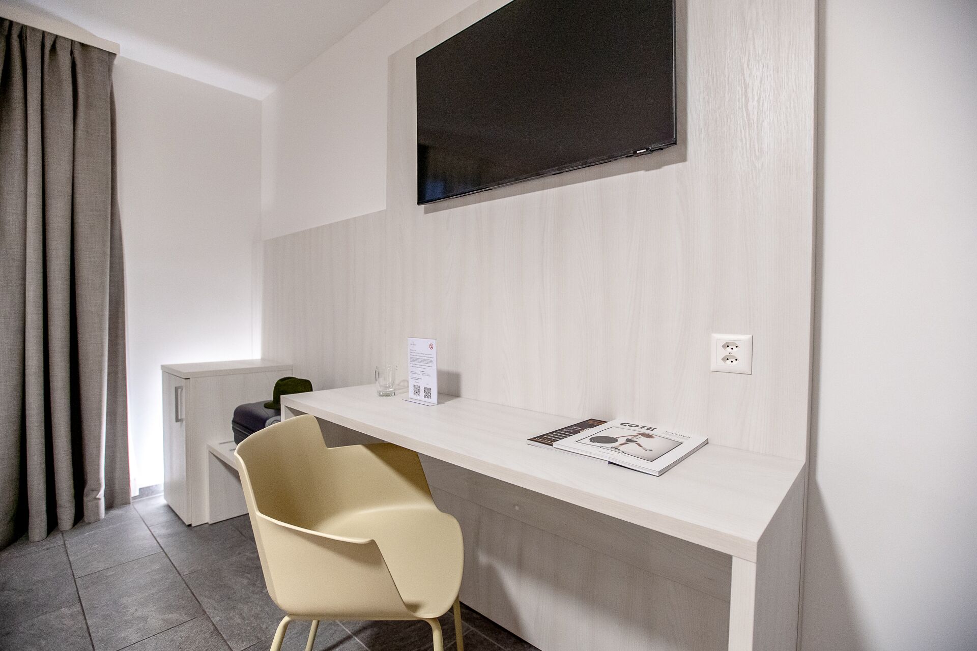 A modern hotel room with a white desk, a flat-screen TV mounted on the wall, a beige chair, and a small table with a magazine.