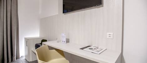Een moderne hotelkamer met een wit bureau, een flatscreen televisie aan de muur, een beige stoel en een klein tafeltje met een tijdschrift.