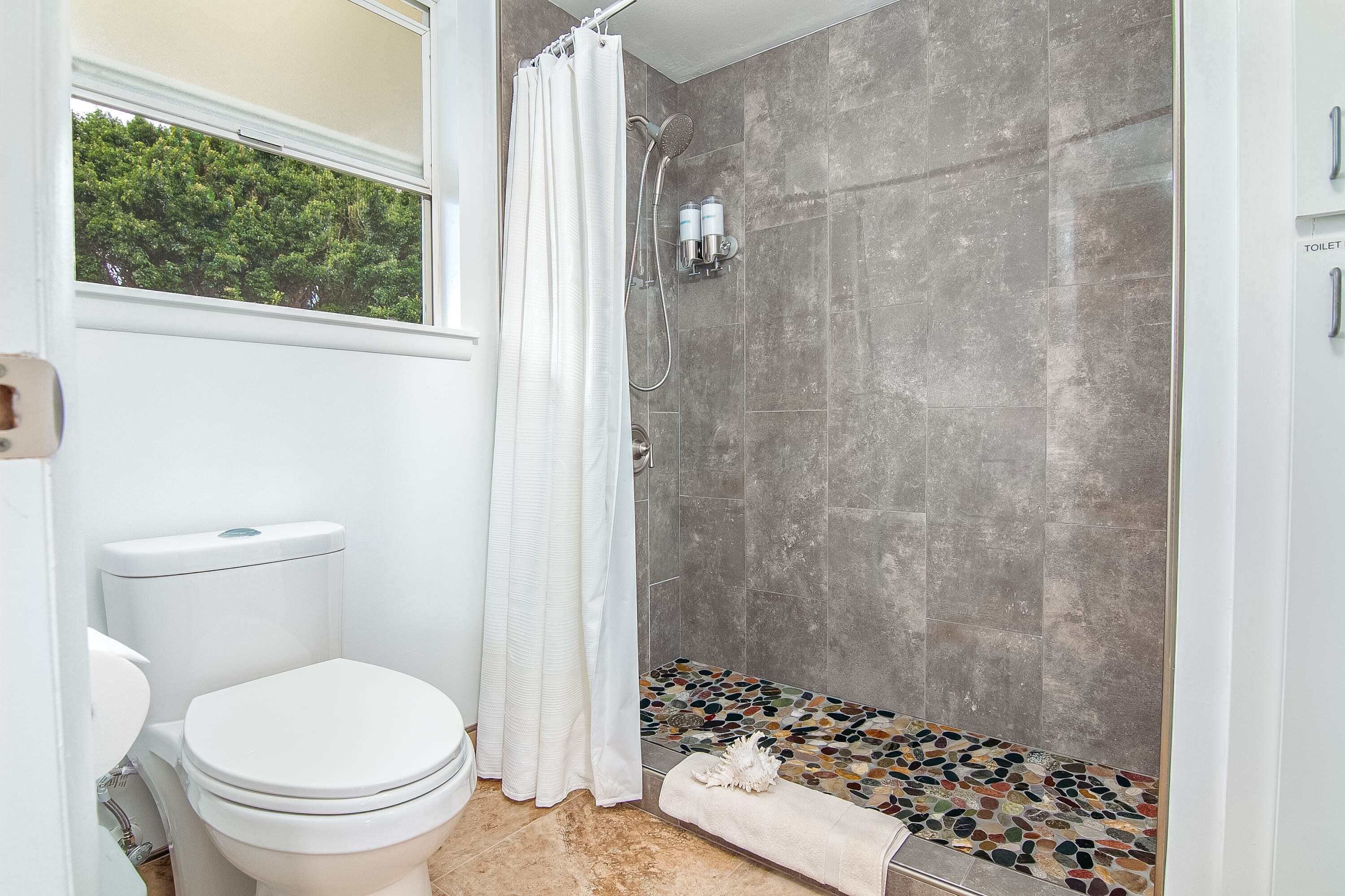 A bathroom with a toilet, shower, and a window with a view of trees.