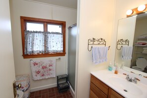A bathroom with a white sink, wooden cabinets, a mirror with lights, a window with curtains, and a shower area. - 36 Salt Marsh Rd (Sandwich)