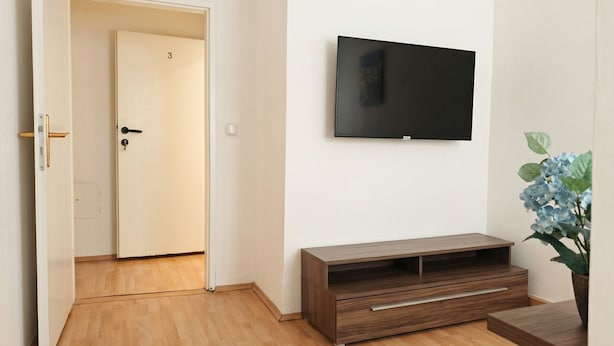 A room with a wooden floor, a TV mounted on the wall, a wooden cabinet, and a door leading to another room. - Big 3BR shared Apt. Perfect location (Vienna)