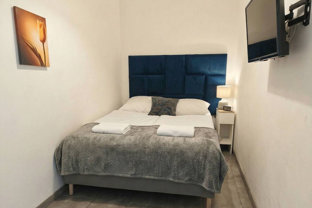 A narrow hotel room with a bed, a nightstand, a TV, and a wall-mounted artwork.