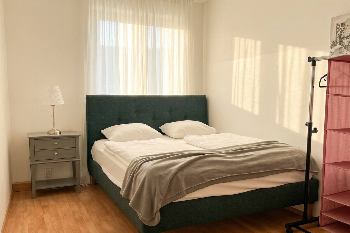 A neatly made bed with a green headboard, a bedside table with a lamp, and a wardrobe with a pull-out shelf.