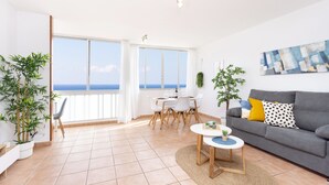 A spacious living room with a grey sofa, a round coffee table, and a dining area with a white table and chairs. There are two large windows offering a view of the sea. - Frontline Duplex Las Arenitas with pool (Candelaria)