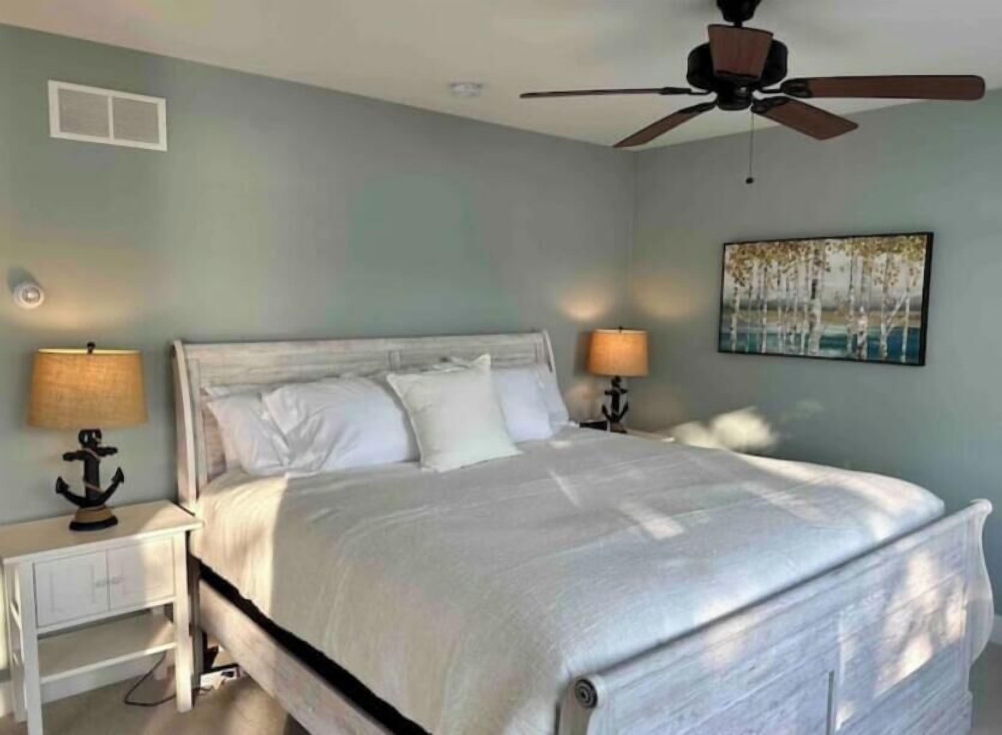 A bedroom with a bed, nightstands, a ceiling fan, and a painting on the wall.
