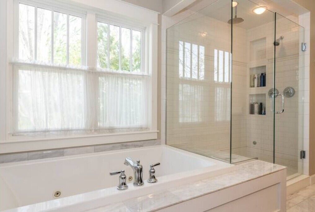 Uma casa de banho moderna com uma banheira grande e branca, um chuveiro com divisória de vidro e uma janela com cortinas translúcidas.