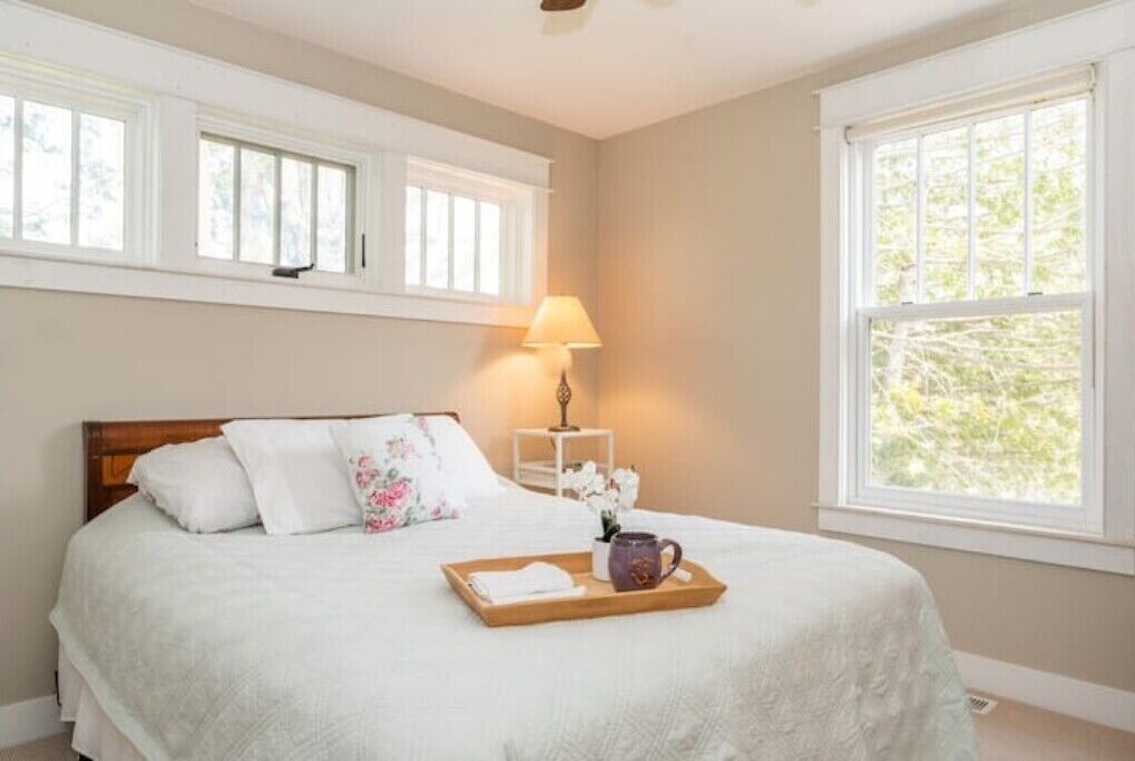 A neatly made bed with a tray of items, a bedside lamp, and a window with a view of greenery.