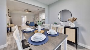 Un coin repas avec une longue table dressée pour quatre personnes, des chaises blanches, un miroir rond et un buffet agrémenté d’objets décoratifs.