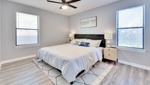 Une chambre à coucher comprenant un lit, des tables de chevet, un ventilateur de plafond et deux fenêtres équipées de stores.