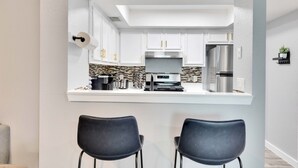 Une cuisine moderne avec des armoires blanches, un réfrigérateur en acier inoxydable et un four encastré. Deux chaises noires sont placées devant le comptoir.