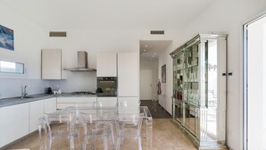 A modern kitchen with a glass dining table and chairs, a built-in oven, and a glass cabinet with decorative items. - Jacuzzi and direct access to the sea, Avola -  DiCasaInSIcilia (Avola)