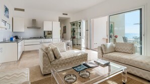 A modern living room with a glass coffee table, two sofas, and a kitchen in the background. - Jacuzzi and direct access to the sea, Avola -  DiCasaInSIcilia (Avola)