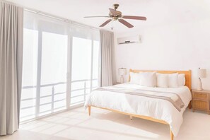 Una habitación de hotel con cama, mesita de noche, ventilador de techo y un amplio ventanal con cortinas.