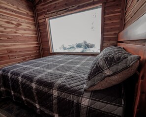 A wooden cabin room with a bed, a window, and a view of a snowy landscape. - Glamping Cabin | Huge Window | Cliff and Animal Views (Green Township)