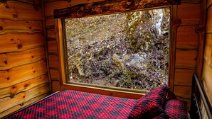 A wooden cabin interior with a bed, a window overlooking a rocky outdoor area.
