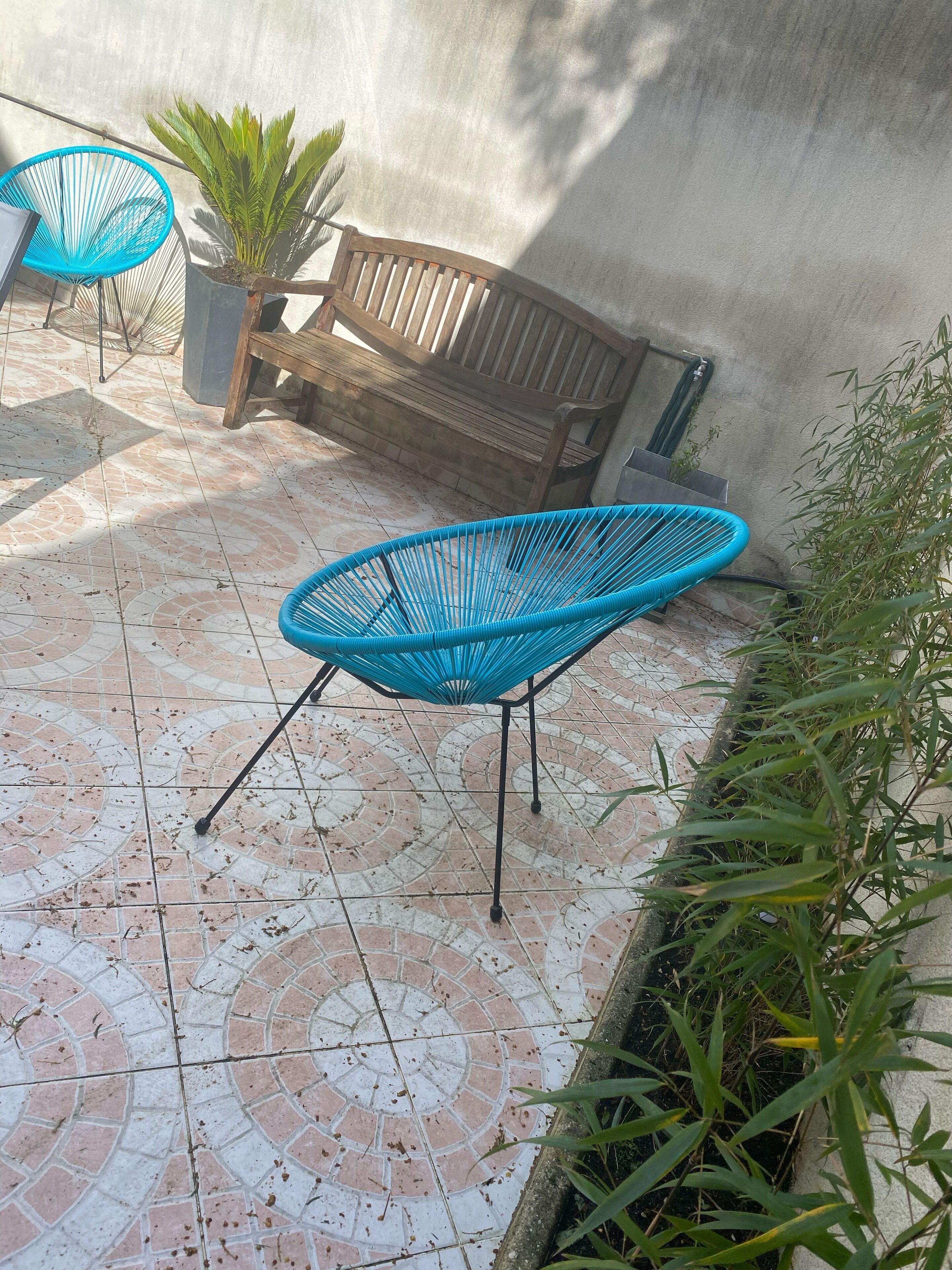 A patio area with a patterned tile floor, a wooden bench, and two blue chairs.