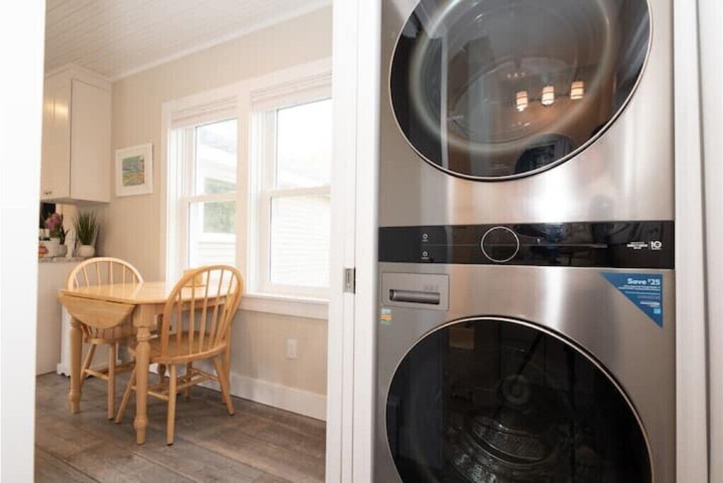 A laundry room with two stacked front-loading washing machines, a wooden dining table with chairs, and a window with blinds.