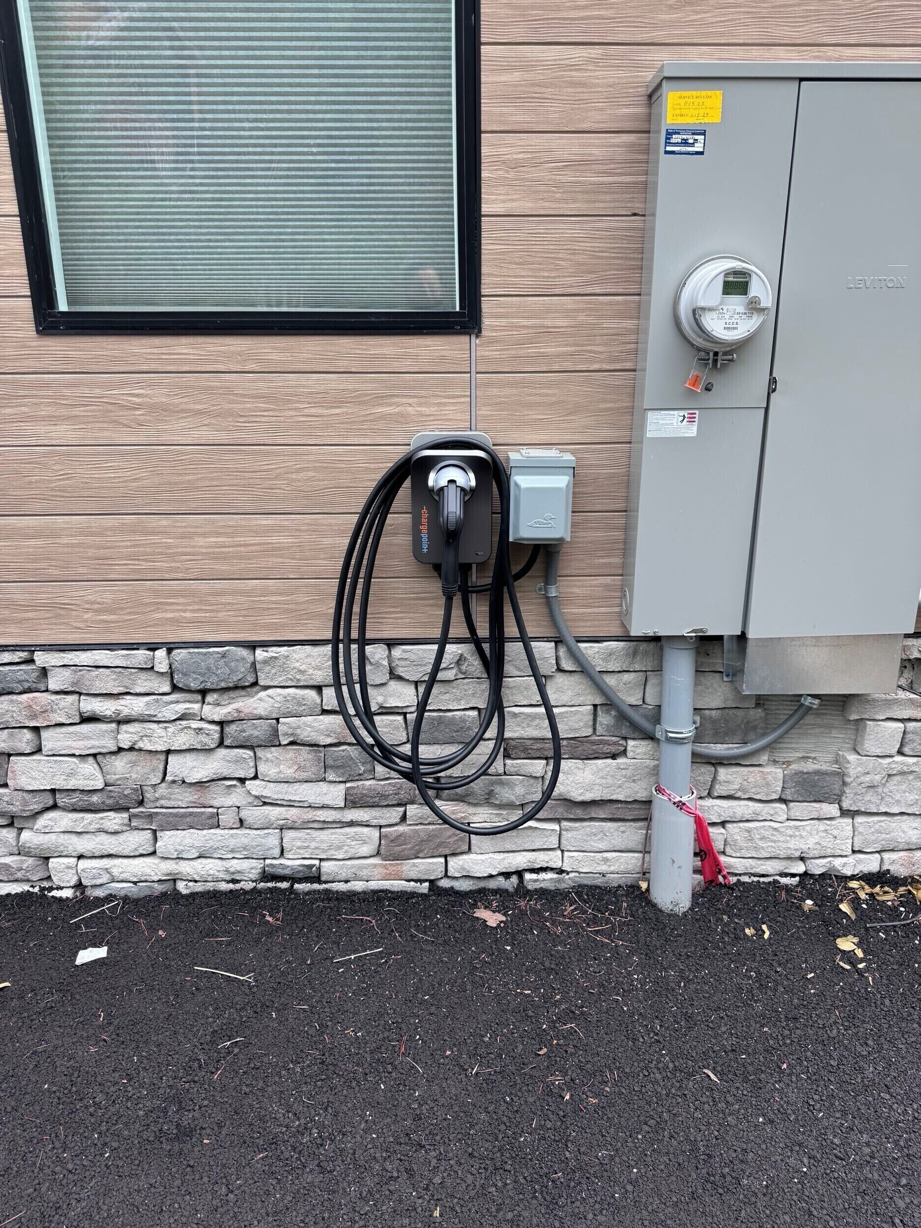 An outdoor electrical box with a meter, connected to a building with a stone foundation and a window with blinds.