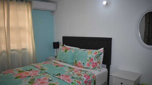 A bedroom with a bed, a bedside table, a lamp, a small drawer unit, and a window with curtains. - Belle Zoon Villa (Gros Islet)