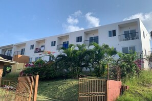 Un immeuble résidentiel à plusieurs étages, doté de balcons, entouré d’une végétation luxuriante et protégé par un portail en métal.