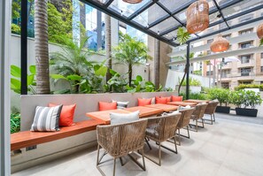 Un espace de restauration en plein air avec des tables en bois et des chaises en osier, entouré d’une végétation luxuriante et doté d’un plafond en verre.