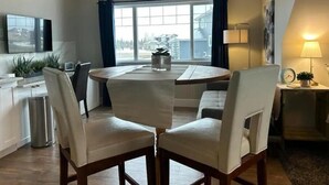 Ein moderner Essbereich mit einem runden Tisch und vier Stühlen, eine Küchenzeile inklusive Spüle und Schränken, sowie ein großes Fenster mit Vorhängen.