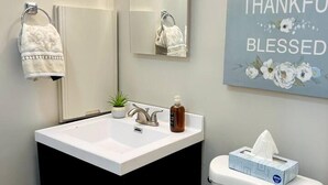 Une salle de bain avec un lavabo blanc, un meuble noir et des toilettes. Un miroir est placé au-dessus du lavabo, et un porte-serviettes est fixé au mur. Un panneau décoratif avec des fleurs et les mots « GRATEFUL THANKFUL BLESSED » est visible.