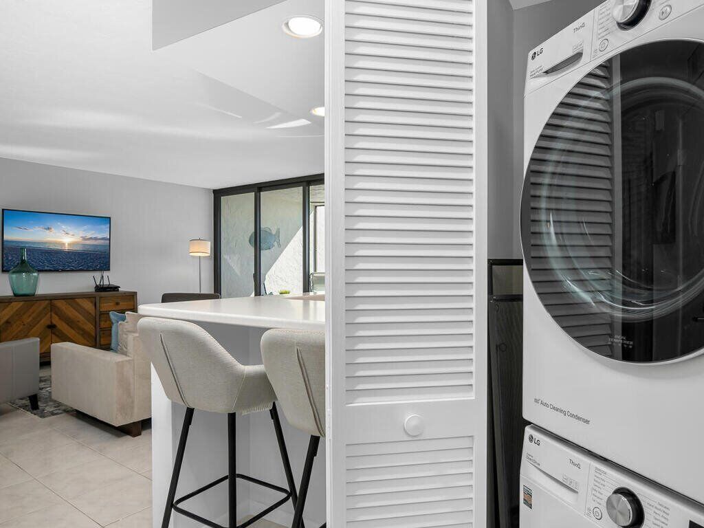 A modern laundry room with a front-loading washer and dryer, a bar area with two stools, and a large window offering a city view.