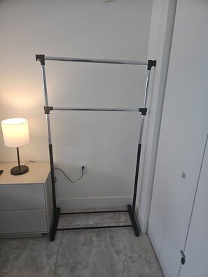 Una habitación con un perchero metálico en la pared, una mesita de noche con una lámpara y un enchufe en la pared.