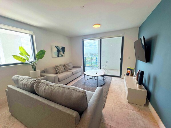 A modern living room with a sofa, a coffee table, a plant, and a balcony with a city view.