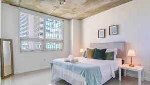 A modern hotel room with a large bed, two bedside lamps, a view of a building through a large window, and a framed picture on the wall. - Stylish 1BR | Free Parking | Walkable Area (Miami)