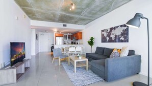 A modern living room with a grey sofa, a coffee table, and a TV on a wooden stand. There is a dining area with chairs and a table, and a kitchen in the background. - Stylish 1BR | Free Parking | Walkable Area (Miami)