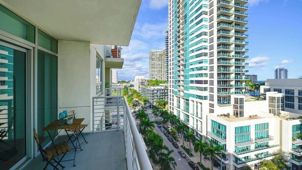 A high-rise building with a balcony featuring a table and chairs, overlooking a street with palm trees and other buildings. - Stylish 1BR | Free Parking | Walkable Area (Miami)