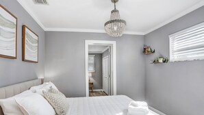 A bedroom with a bed, a chandelier, a door leading to another room, and a window with blinds. - Golf and Pool Vacation Home!
 (St. Petersburg)