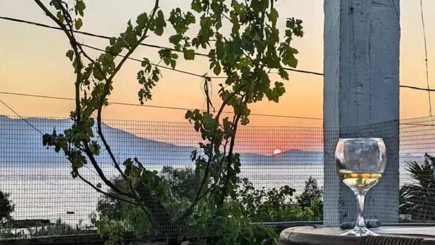 Une table avec un verre de vin blanc, une vue sur un coucher de soleil se reflétant sur les montagnes et une clôture en treillis métallique.