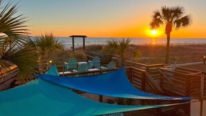 Eine Terrasse am Strand mit Adirondack-St ühlen, einem Holzdeck und Blick auf den Sonnenuntergang über dem Meer.