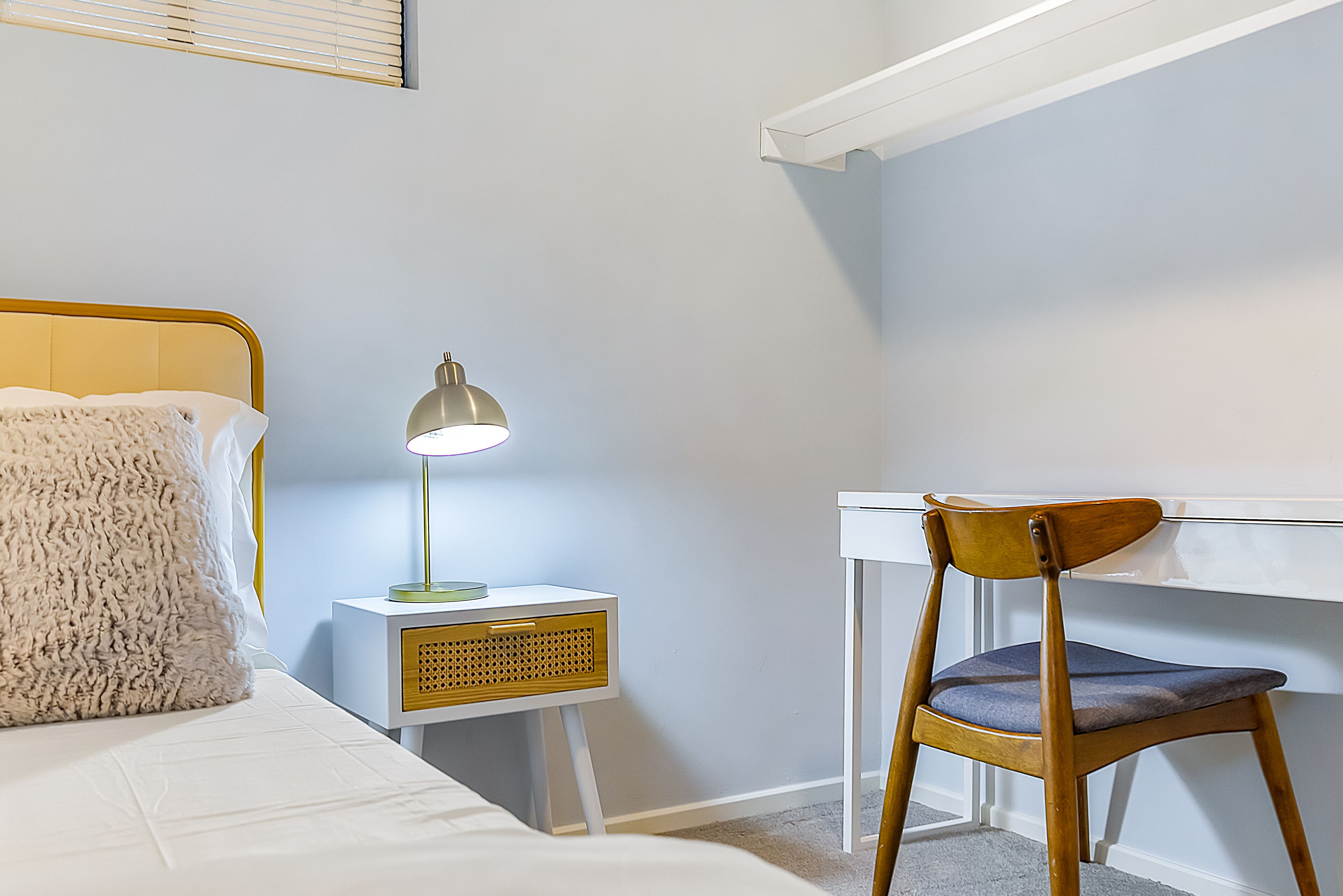 A bedroom with a bed, a bedside table with a lamp, a chair, and a desk.