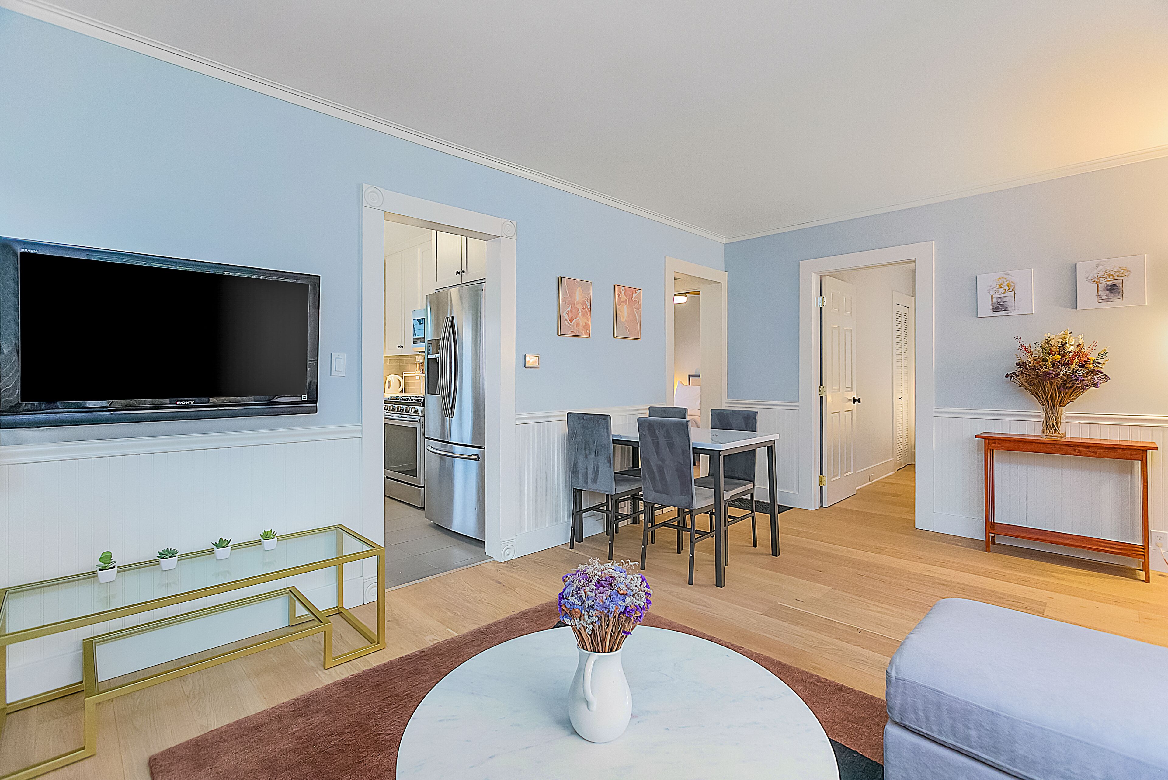 A modern living room with a flat-screen TV, a dining area with chairs and a table, and a kitchen in the background.